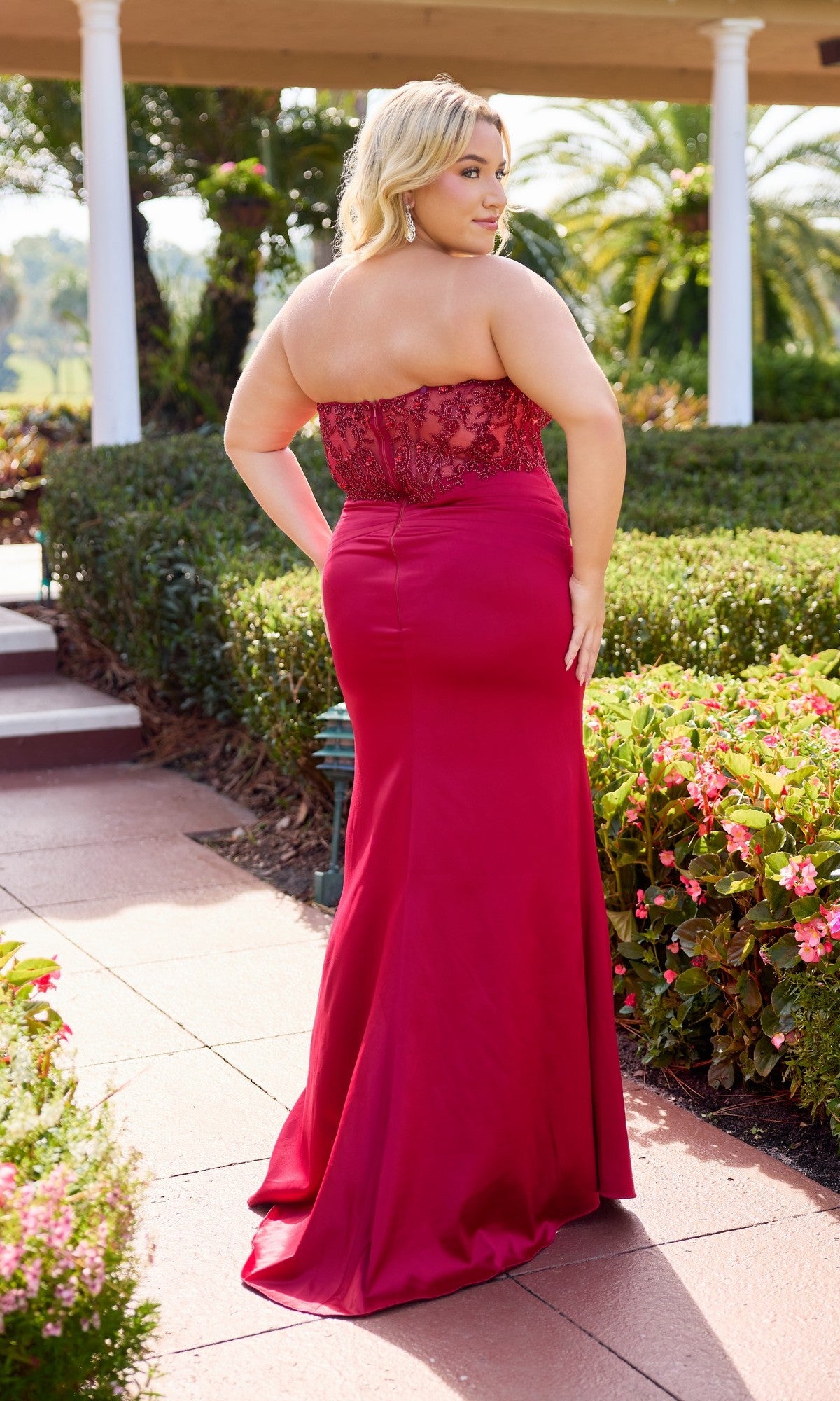 A woman stands outdoors among greenery and flowers, wearing the Long Plus-Size Prom Dress: Primavera 14109, which features a strapless sheer embroidered bodice and a satin sheath skirt.